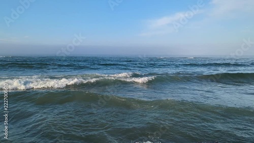 Sea waves roll and splash white foam and water droplets under blue hazy sky