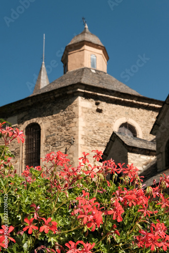 Red geranium flowers (in focus) blooming in front of the historic Romanesque Church of Sant Feliu de Vilac (out of focus) on sunny summer day. Aran Valley, Spain.