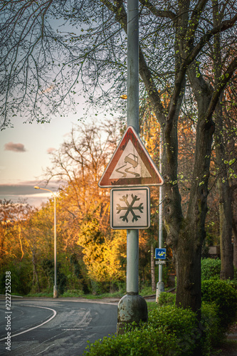 Triangular slippery road traffic sign with snowflake warning sign framed by autumn trees