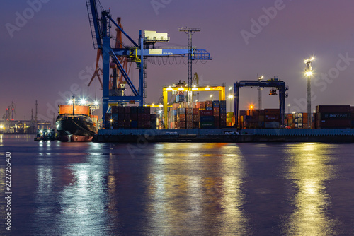 Nighttime port with towering cranes and glowing lights. Container ships float beneath artificial illumination. Water reflects lights, creating shimmering patterns. Heavy machinery stands still
