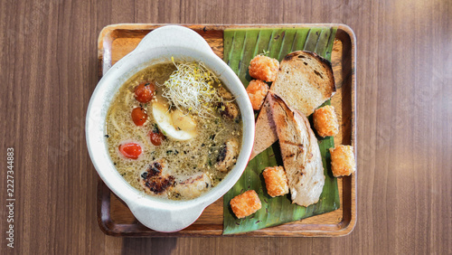 A bowl of hearty chicken soup with grilled chicken, cherry tomatoes, sprouts and egg, served with toasted bread and crispy chicken bites on a wooden tray lined with banana leaf.