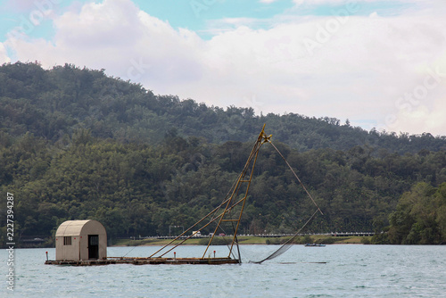 Rustic Fishing Hut and Net Platform Floating on Calm Lake with Forested Mountains, Rural Asian Fishery