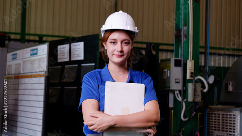 A female engineer wearing a white safety helmet and blue work uniform holds documents inside an industrial factory. She displays a confident and professional attitude, surrounded by machinery, steel s