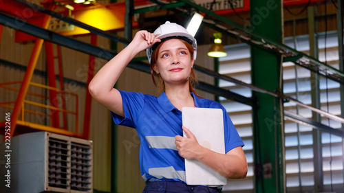 A female engineer wearing a white safety helmet and blue uniform holds documents and reviews information confidently inside an industrial factory.