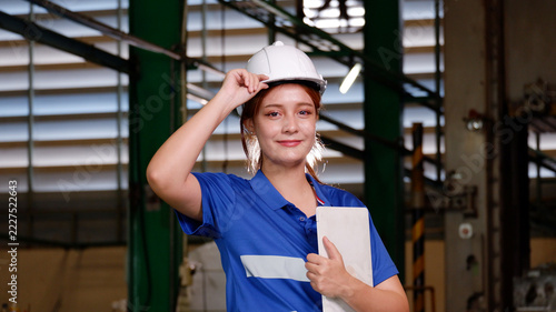 A female engineer wearing a white safety helmet and blue work uniform holds documents inside an industrial factory. She displays a confident and professional attitude, surrounded by machinery, steel s
