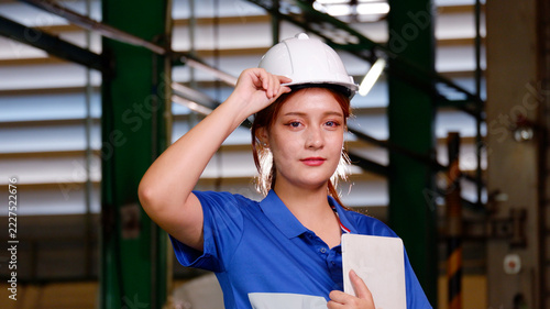 A female engineer wearing a white safety helmet and blue work uniform holds documents inside an industrial factory. She displays a confident and professional attitude, surrounded by machinery, steel s