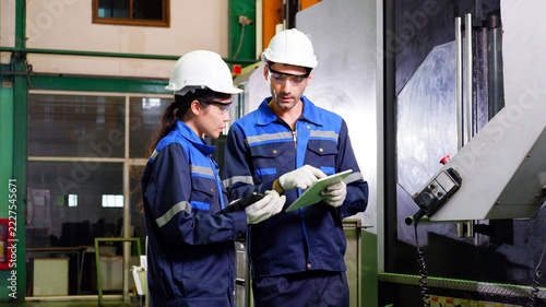 Male and female engineers collaborate to inspect machine components and record operational data inside an industrial factory.