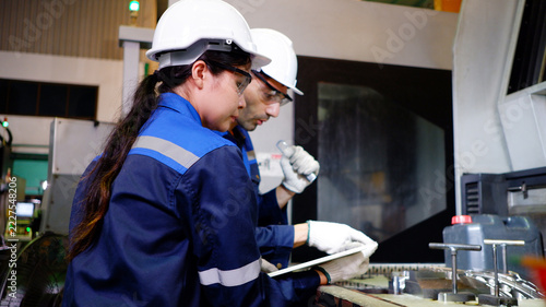 Male and female engineers collaborate to inspect machine components and record operational data inside an industrial factory.