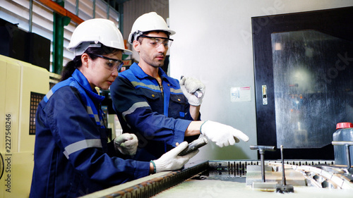 Male and female engineers collaborate to inspect machine components and record operational data inside an industrial factory.