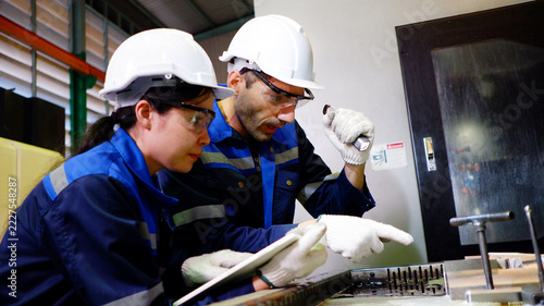 Male and female engineers collaborate to inspect machine components and record operational data inside an industrial factory.