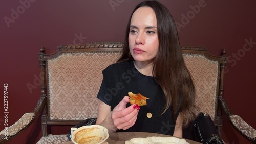 Young brunette woman chewing a croissant with fish and vegetables while holding a piece at a table indoors, casual dining, savory snack