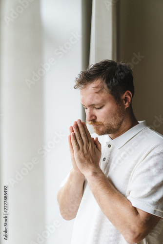 Christian man praying with clasped hands by window