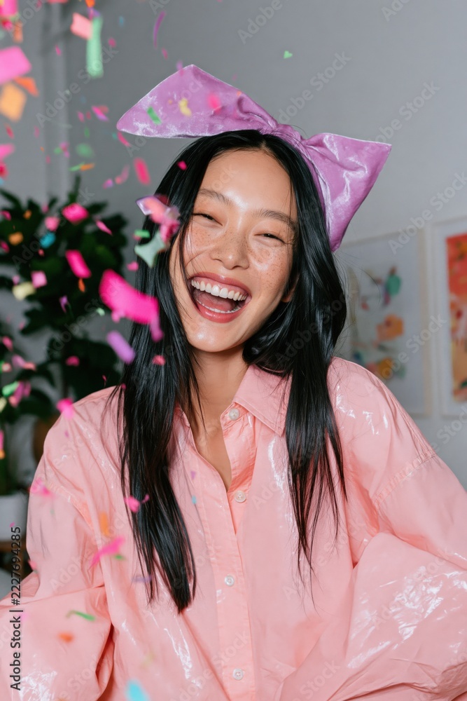Fototapeta premium Joyful young woman laughing with confetti and a large pink bow for party poster and celebration background with playful vibes