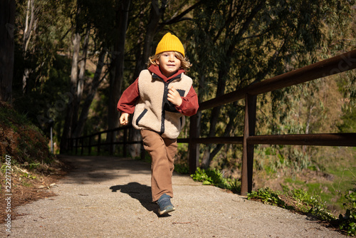 Young child running on forest path with wooden railing, wearing yellow hat and warm clothes, active outdoor lifestyle, motion, childhood adventure concept