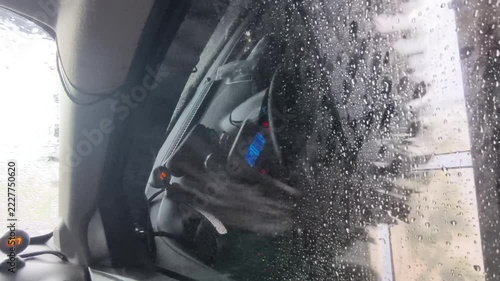 Inside view of car driving through automated car wash machine with water, soap foam and rotating brushes on windshield
