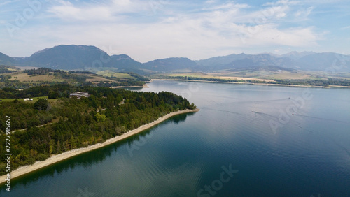 Majestic aerial view of tranquil blue water reservoir surrounded by lush green forests and towering mountain peaks under a dramatic cloudy sky inviting wanderlust and peaceful outdoor escape, ‎⁨Liptov