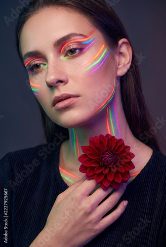Young woman with creative neon light makeup holding red flower in studio portrait. Concept of rainbow makeup, beauty portrait.