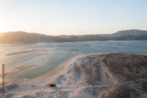 Aerial view of sand dunes and a wooden boardwalk leading to a beach bar on Troia, Portugal. The beach borders a calm lagoon with sandbars in warm evening light.