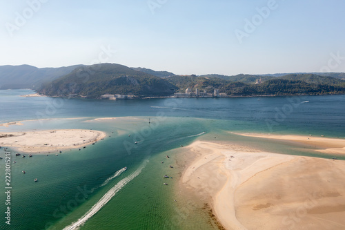 Aerial view of a boat leaving a white wake between sandbars at the mouth of the Sado estuary near Troia, Portugal. Anchored boats line the sand below Arrabida.