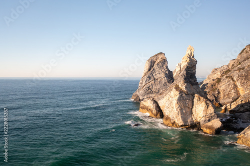 Aerial view of jagged sea stacks rising from the Atlantic Ocean at Ursa Beach near Sintra, Portugal. Warm evening sun lights the rocks, with clear sky on the left.