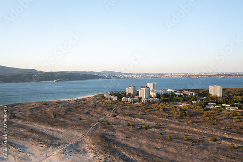 Aerial view of white resort towers among pines on Troia, Portugal, at golden hour. Dunes with a boardwalk fill the foreground, with Setubal across the estuary.