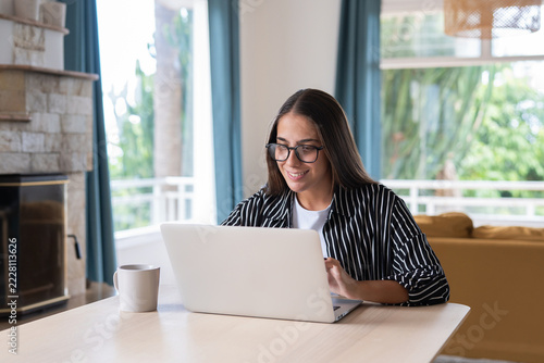 Obraz na plátně Woman plans a trip on a laptop beside a mug in her living room, smiling as she researches travel