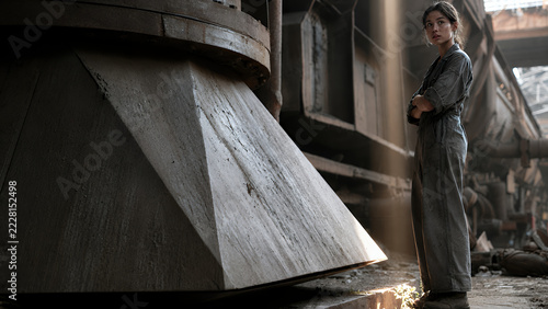 Young woman worker in a grey boiler suit standing next to massive metallic industrial structures in a factory setting