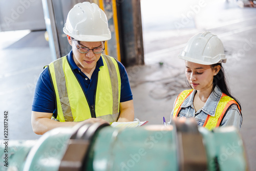 Two workers in safety vests and hard hats review a checklist beside industrial machinery in a factory, focusing on maintenance and safety procedures.