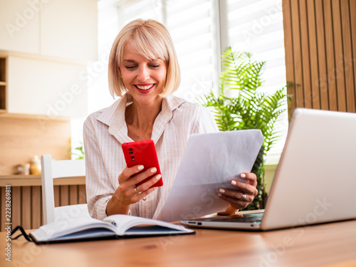 Cheerful woman reviewing financial paperwork while multitasking with digital devices from home.