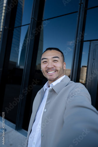 Man taking a smiling selfie outdoors by urban glass building