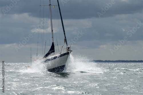 Segelyacht durchquert aufgewühlte Küstengewässer und erzeugt eine mächtige Gischtwand unter dramatischen grauen Wolken bei kühlem, kontraststarkem Tageslicht.