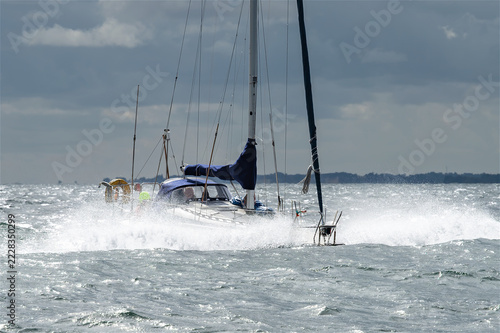 Segelboot kämpft sich durch kabbelige See, während kräftiger Wind hohe Gischt aufwirbelt und silberne Sonnenreflexe die dynamische maritime Szene unter bewölktem Himmel beleuchten.