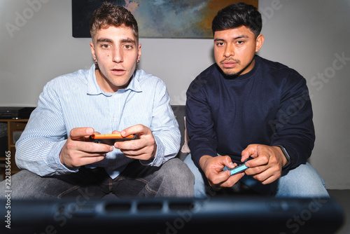 Two young men sitting on a couch in a living room, playing a video game together. They hold handheld controllers and watch the screen with focused and surprised expressions.