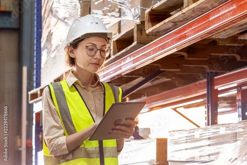 Female warehouse supervisor using a digital tablet for inventory tracking. Focused woman logistics worker in white hard hat and safety vest. Smart supply chain operator managing orders on touchscreen.