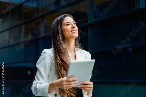 Young professional woman holding tablet outside modern office building smiling confidently and gazin