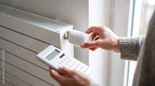 Close-up of hand adjusting white radiator thermostat valve while holding calculator indoors. Concept of heating cost calculation, energy saving, and utility bill management