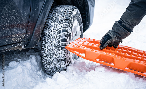 Close-up of hand in glove placing orange traction track under car tire stuck in deep snow. Concept of winter driving rescue, off-road recovery, and vehicle emergency assistance