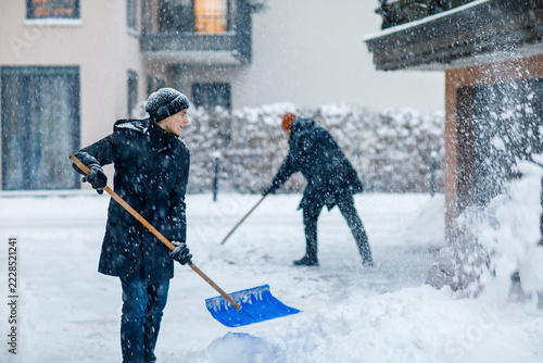 Teenage boy and father shoveling snow together in a residential courtyard, winter clothing, snowfall, everyday family life outdoors.