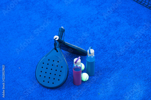 Dark padel paddles and neon tennis balls are resting on bright blue court with colorful bottles