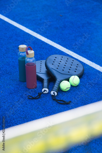 Two black padel paddles are lying on blue court near net, neon-yellow balls and bottles nearby
