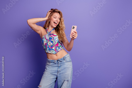 Happy young woman taking a selfie with smartphone against purple background wearing colorful halter top and ripped jeans, casual summer lifestyle and joyful expression
