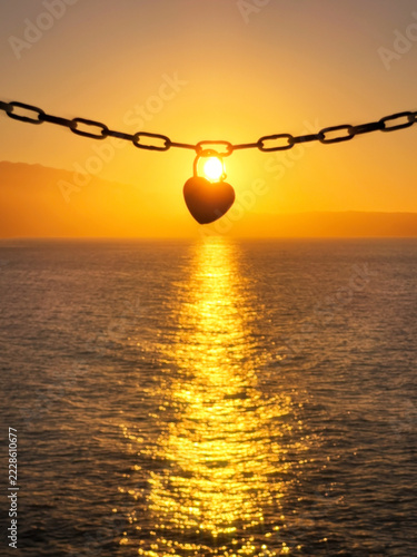 Romantic heart-shaped padlock hanging from a metal chain with the setting sun shining through it over a calm sea, symbolizing love, commitment, romance, and lasting relationships.