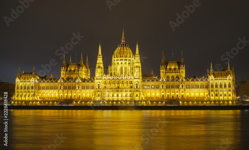 View of the illuminated Hungarian Parliament Building beside the Danube River in Budapest, Hungary, at night, with golden reflections and its historic Gothic Revival facade in winter.