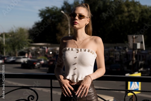 Half body portrait of a young caucasian woman in sunglasses and white top. She poses with hands on a belt and brown leather pants outdoors in dramatic sunlight.