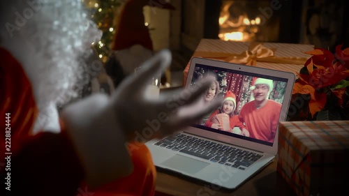 Santa Claus video calling family with little girl on laptop, greeting child by webcam, opening Christmas gift box in virtual online meeting using computer sitting in his decoration residence with
