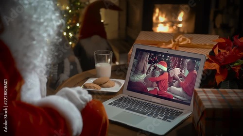 Santa Claus video calling family with little girl on laptop, greeting child by webcam, opening Christmas gift box in virtual online meeting using computer sitting in his decoration residence with