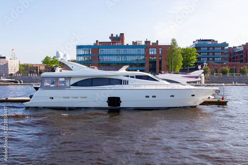 White yacht glides calmly on urban waterway. Modern buildings loom in soft daylight behind. Sunlight reflects off clean, polished hull. Calm waters ripple gently around vessel