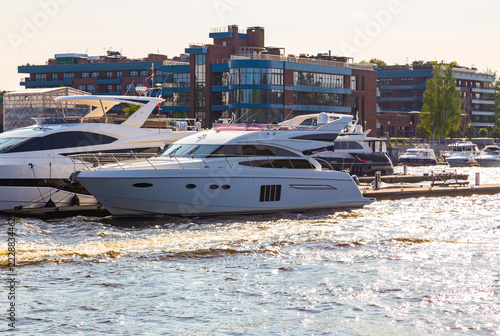 Modern yachts glide through calm waters near urban docks. Sunlight glints off their sleek, polished hulls. Cityscape looms behind, blending glass and brick