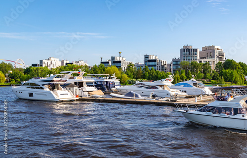 Sunny day glows over tranquil harbor. Yachts float peacefully on calm waters. Modern buildings stand tall behind green trees. People relax on boats, enjoying leisure time. Perfect for vacation, luxury