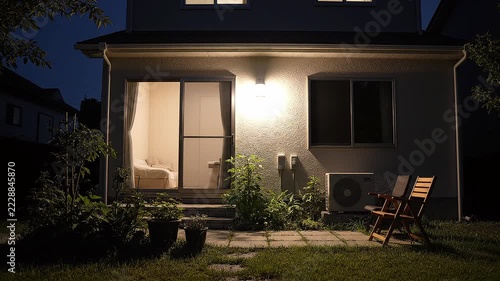 A wall light switches on at night, illuminating the entrance, garden path, plants and patio of a modern home. This fixed wide shot captures warm exterior lighting against the dark blue evening sky.